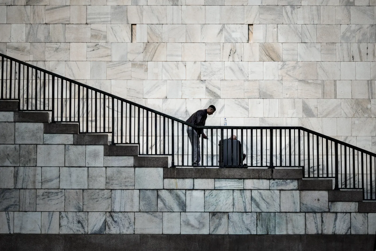 Mann steht mit Koffer auf breiter Treppe vor Marmormauer